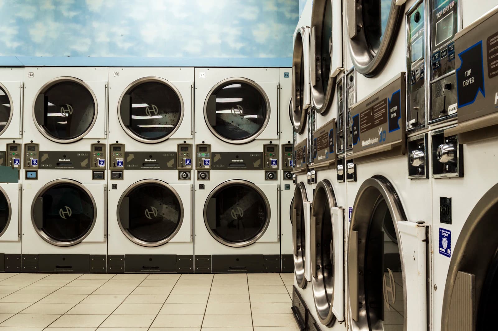 Rows of white commercial laundry machines beside an open tiled floor and a soft blue wall